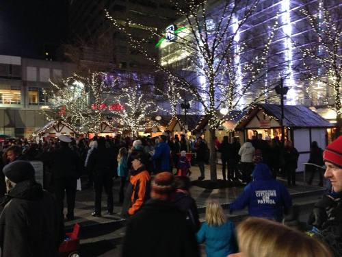 A bustling night scene with crowds, festive lights, and holiday market stalls in a city square.