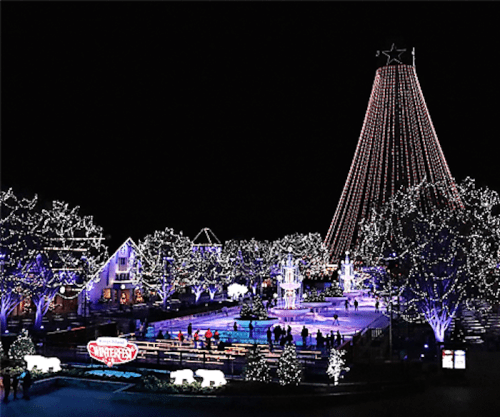 A festive winter scene with a large lit Christmas tree, ice skating rink, and illuminated trees at night.