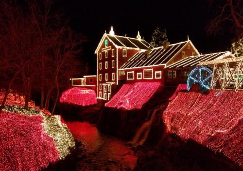 A beautifully lit house surrounded by vibrant holiday lights, reflecting in a nearby stream at night.