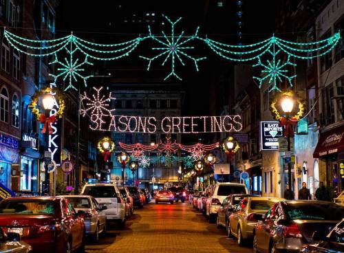 A festive street decorated with colorful lights and a "Seasons Greetings" sign, lined with parked cars at night.