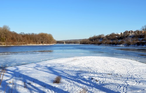 A snowy riverbank under a clear blue sky, with ice on the water and trees lining the shore.