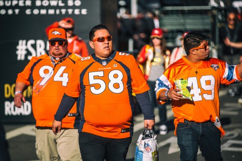 Three fans in orange Denver Broncos jerseys walk together, enjoying the Super Bowl atmosphere.