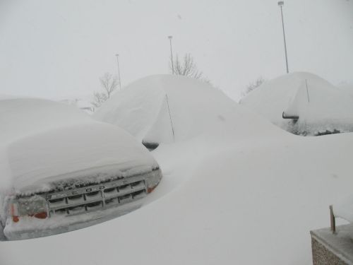 Cars buried under heavy snow in a winter storm, with only their roofs visible above the snowdrifts.