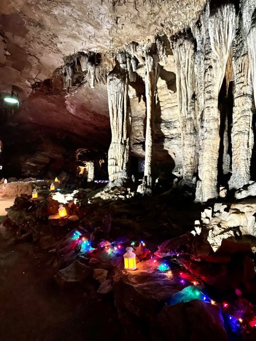 Stalactites and stalagmites in a cave, illuminated by colorful lights and lanterns along the rocky path.