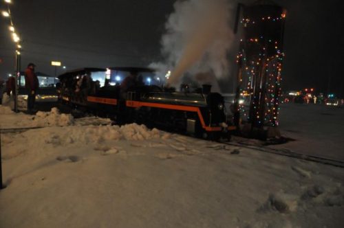 A steam train decorated with lights, releasing smoke, parked in a snowy landscape at night with people nearby.