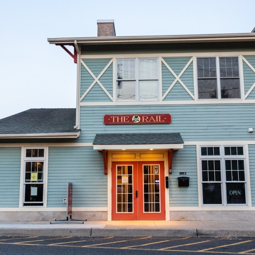 Exterior of a blue building with a red sign reading "The Rail" and orange double doors, illuminated at dusk.