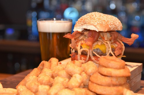 A towering burger with bacon and cheese, served with crispy onion rings and a glass of beer in the background.
