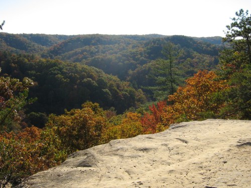 A scenic view of rolling hills covered in autumn foliage, with a rocky foreground and bright sunlight.