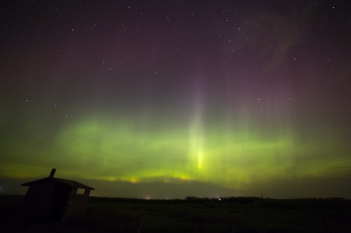 A vibrant display of the northern lights illuminates the night sky above a silhouette of a small shed.