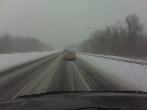 A snowy highway scene with limited visibility, featuring a car driving on a snow-covered road.