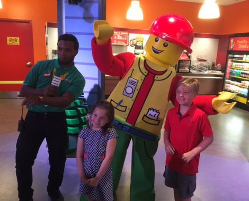 A costumed LEGO character poses with two children and an adult in a colorful indoor setting.