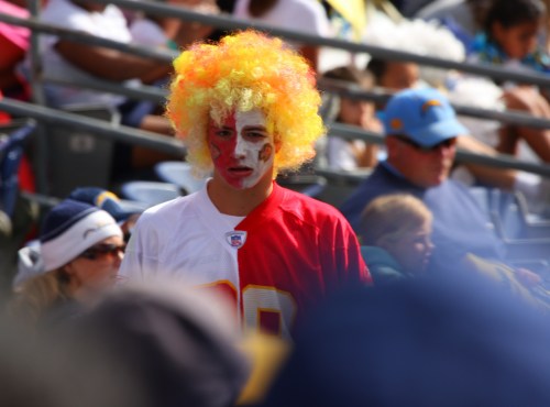 A fan with a bright yellow and orange clown wig and face paint, wearing a red jersey, stands in a crowded stadium.