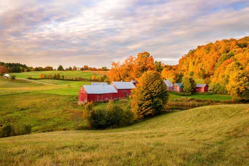 A scenic autumn landscape featuring red barns, colorful trees, and rolling green hills under a cloudy sky.