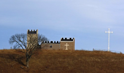 A hilltop scene featuring a building with a cross design and a large white cross against a blue sky.