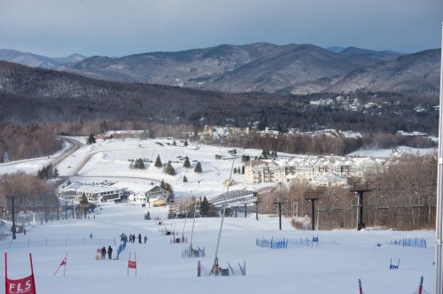 A snowy ski slope with skiers, overlooking a mountain landscape and a small town in the distance.