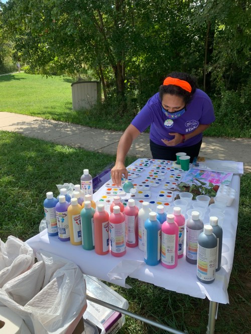 A person in a purple shirt arranges colorful paint bottles on a table outdoors, preparing for an art activity.