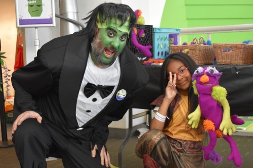 A man in a Frankenstein costume poses with a girl holding a colorful puppet, both smiling and having fun.