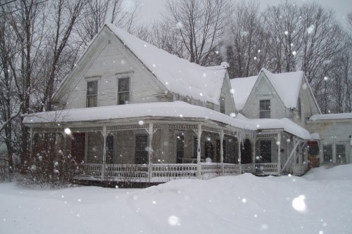 A snow-covered house with a large porch, surrounded by trees, during a snowfall.