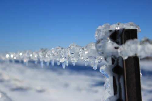 Icicles hang from a black fence against a clear blue sky, with a snowy landscape in the background.
