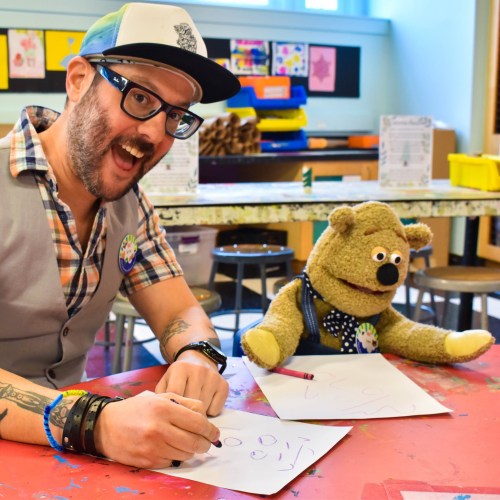A smiling man with glasses and a cap sits at a table, drawing with a puppet beside him in a colorful classroom.