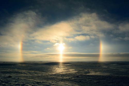 A serene landscape featuring a bright sun with two sun dogs and a cloudy sky, reflecting on a snowy ground.