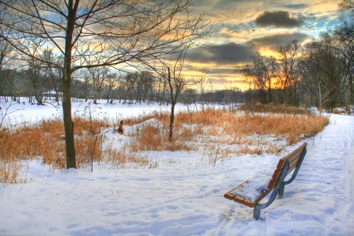 A snowy landscape at sunset, featuring a bench beside a frozen field and bare trees against a colorful sky.