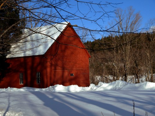 A red barn surrounded by snow, with trees and a clear blue sky in the background. Shadows stretch across the snow.