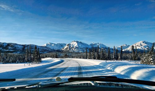 A snowy road winds through a winter landscape with mountains and evergreen trees under a clear blue sky.