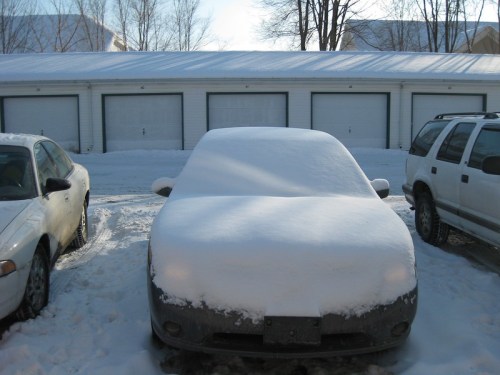 A car covered in snow parked in front of garages, with sunlight shining on the snowy scene.