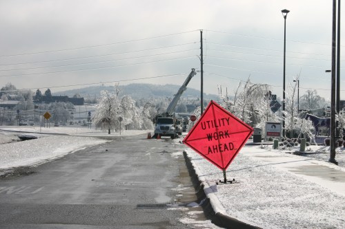 A snowy road with a bright orange sign reading "Utility Work Ahead" and a utility truck in the background.