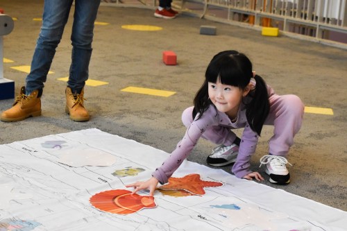 A young girl in purple clothes points at a colorful sea creature on a large map while playing on a carpeted floor.