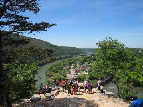 A scenic view from a mountain overlook, showing a river, lush greenery, and a group of hikers enjoying the landscape.