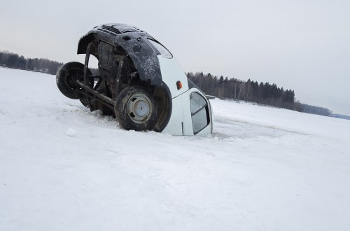 A white car is partially submerged in snow and ice, overturned on a frozen landscape with trees in the background.