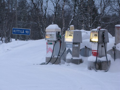 Snow-covered gas pumps beside a road sign for Kittilä, with trees in the background and a wintery landscape.