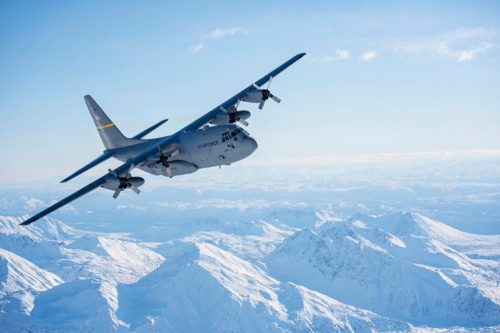 A military aircraft flying over snow-capped mountains under a clear blue sky.