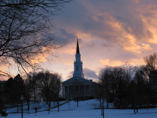 A church with a tall steeple stands against a colorful sunset, surrounded by bare trees and snow-covered ground.