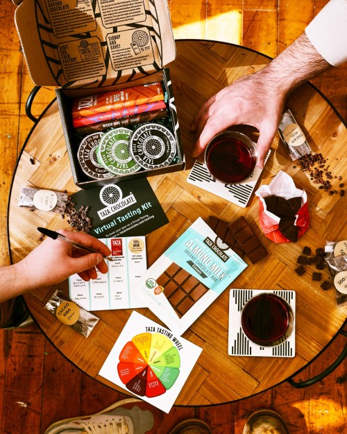 A table with a chocolate tasting kit, tasting notes, chocolate bars, and two hands holding cups and a pen.