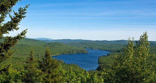 A serene landscape featuring a blue lake surrounded by lush green forests and distant mountains under a clear sky.