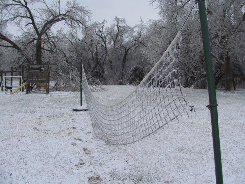 A snowy landscape featuring a frozen volleyball net and a playground in the background, surrounded by icy trees.