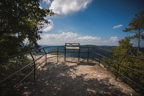 A scenic overlook with a railing, featuring a sign and panoramic views of mountains and a blue sky with clouds.
