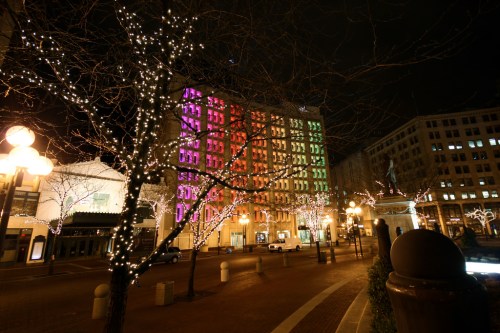 A brightly lit building at night, adorned with colorful lights, surrounded by trees decorated with white lights.