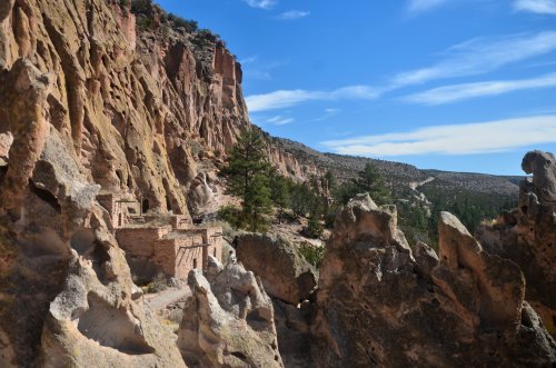 Rocky cliffs and formations surround ancient structures nestled among trees under a clear blue sky.