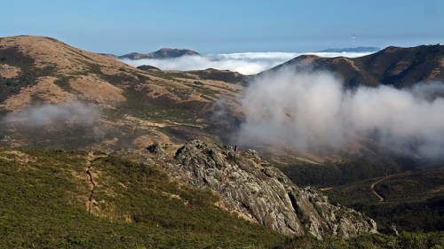A scenic view of rolling hills and mountains, with clouds hovering over the landscape under a clear blue sky.