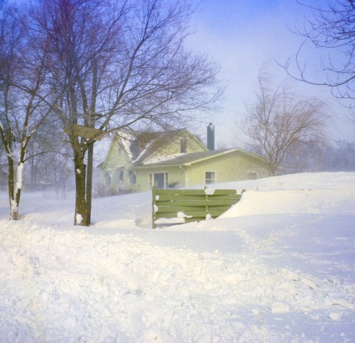 A snow-covered house surrounded by bare trees in a winter landscape, with a clear blue sky above.