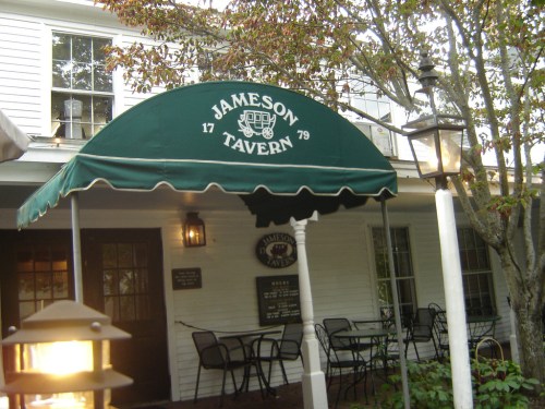 Green awning with "Jameson Tavern 1779" over the entrance of a historic building, surrounded by trees and outdoor seating.