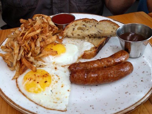A plate with two fried eggs, sausage links, crispy fried onions, toast, and dipping sauces.