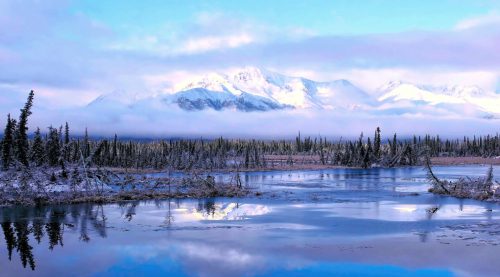 A serene landscape featuring a calm lake, snow-capped mountains, and misty clouds reflecting in the water.