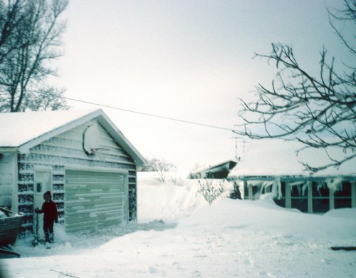 A child stands beside a snow-covered garage, surrounded by deep snow and bare trees in a winter landscape.
