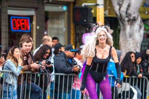 A lively parade scene with a woman in a black outfit and colorful accessories, surrounded by onlookers and bubbles.