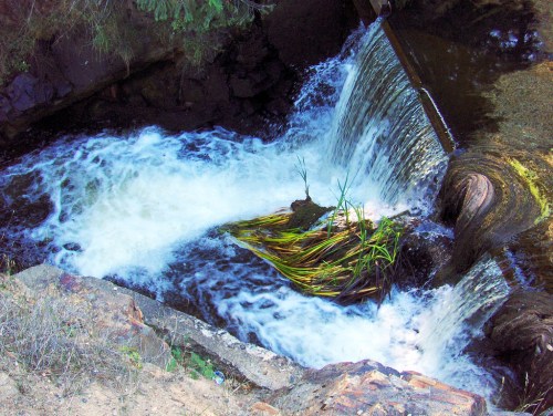 A small waterfall cascades over rocks into a flowing river, surrounded by greenery and natural vegetation.